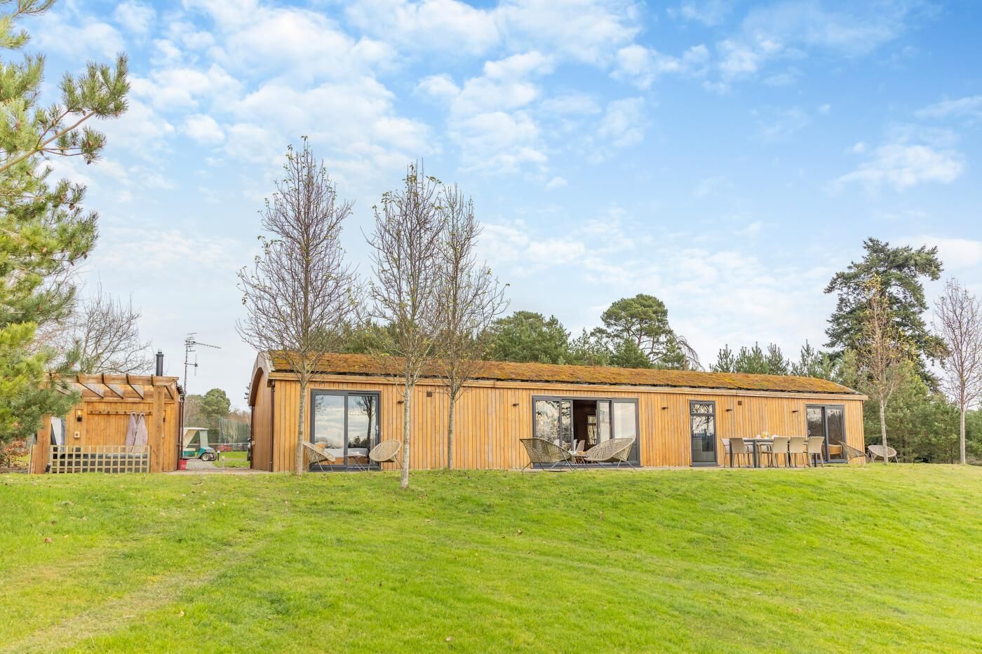 Una cabaña moderna de madera con un amplio ventanal corredero, un patio con mobiliario exterior y una pequeña construcción adyacente.