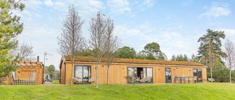 Una cabaña moderna de madera con un amplio ventanal corredero, un patio con mobiliario exterior y una pequeña construcción adyacente.