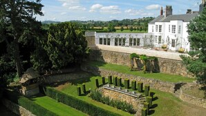 A landscaped garden with a stone wall, a white building with multiple windows, and a view of the countryside.