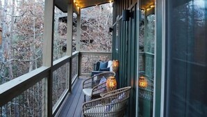 Eine Holzterrasse mit Lichterketten, Korbstühlen und einem Glasgeländer mit Blick auf einen bewaldeten Bereich.