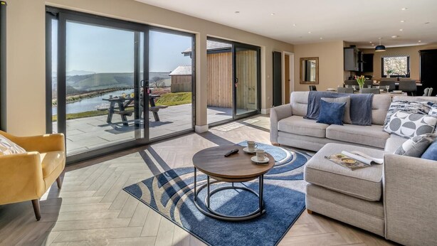 A spacious living room with a large sliding glass door leading to an outdoor patio, a sofa set, a coffee table, and a dining area visible in the background. - 3 Bed in Merthyr Cynog (oc-g30312) (Brecon)
