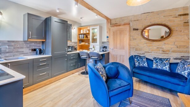 A modern kitchen with grey cabinets, a wooden ceiling, and a blue sofa with patterned cushions. - 1 Bed in Skipton (oc-w30426) (Skipton)