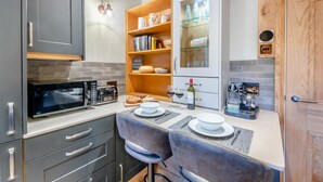 A compact kitchen with a microwave, toaster, and coffee machine. There are two bar stools and a small dining table with plates and glasses. - 1 Bed in Skipton (oc-w30426) (Skipton)