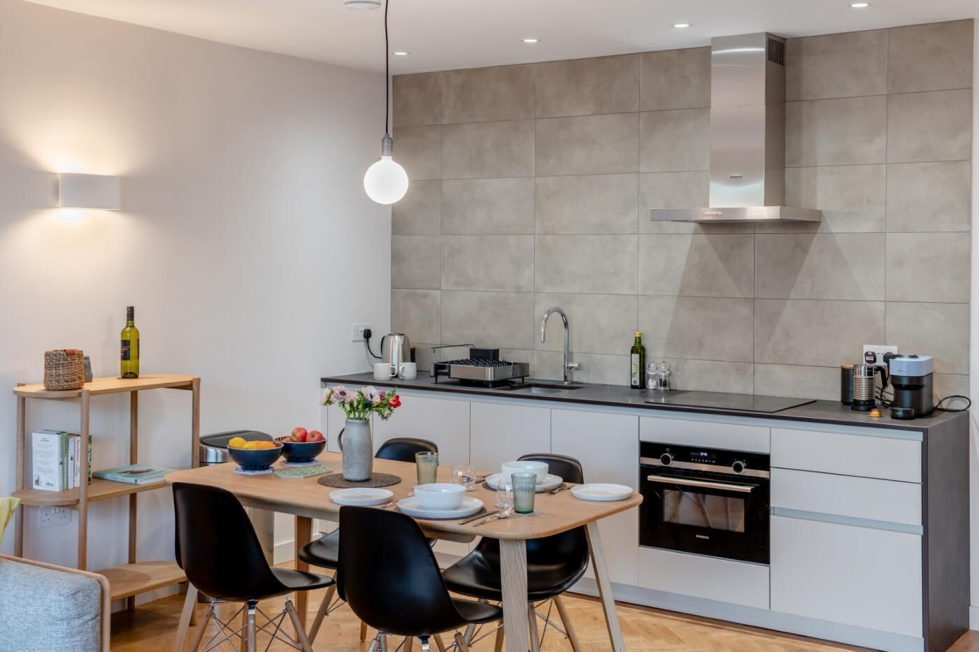 A modern kitchen with a dining area, featuring a built-in oven, coffee machine, and a hanging light fixture.