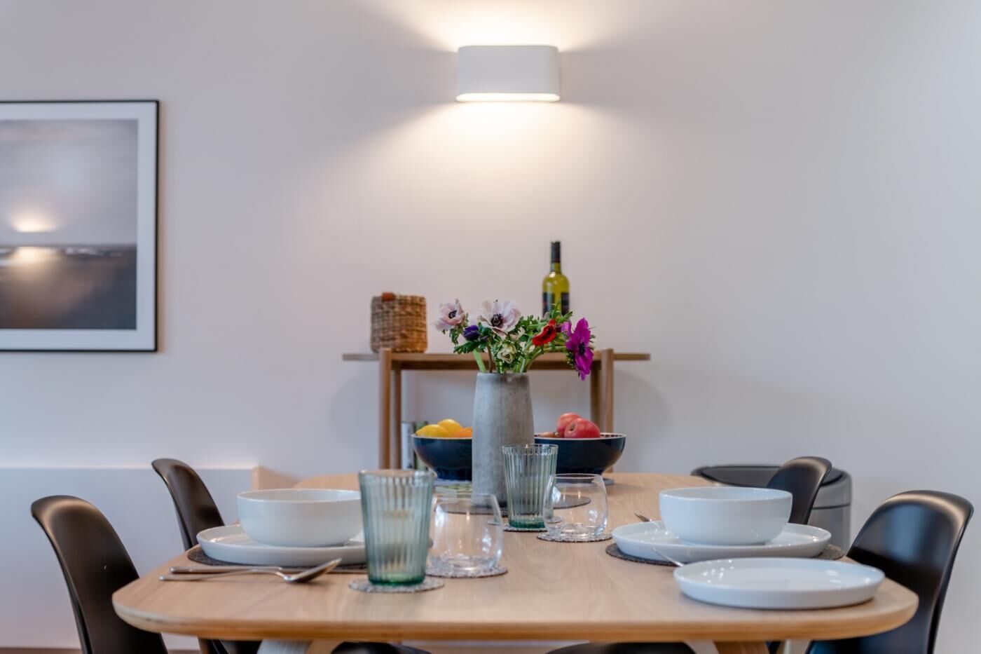 A dining table set with white plates, glasses, and cutlery, a vase with flowers, and a bottle on a shelf.