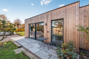 A modern wooden house with large glass doors, a patio with a table and chairs, and a car parked nearby. - 1 Bed in Iden (oc-d30377) (Iden)