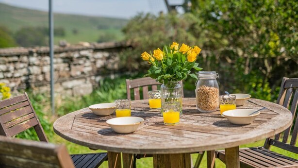 A round wooden table set outdoors with a vase of yellow flowers, a glass jar, and bowls with orange juice. - 3 Bed in Hawes (oc-s30429) (Hawes)