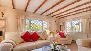 Ein gemütliches Wohnzimmer mit einer Couch, Sesseln und einem Couchtisch. Es gibt Fenster mit Vorhängen, eine Lampe und eine Vase mit Blumen.