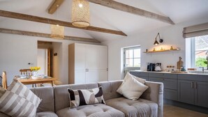 A modern living room with a grey sectional sofa, wooden beams on the ceiling, and a kitchen area with grey cabinets and a window. - 3 Bed in Whitwell (oc-d30478) (Godshill)