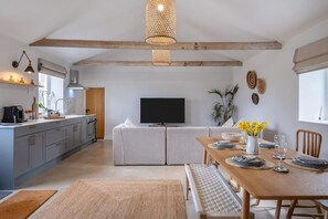A modern kitchen with a dining area, wooden beams, a flat-screen TV, and a dining table set for four. - 3 Bed in Whitwell (oc-d30478) (Godshill)