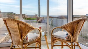 Two wicker chairs with cushions, a small table with a cup and saucer, and a view of a coastal area through large windows. - 2 Bed in Beadnell (oc-f30468) (Beadnell)