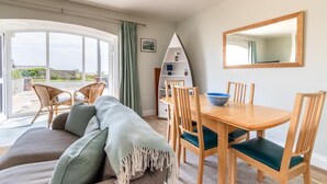 A cozy living room with a sofa, dining table, and chairs, a bookshelf, and a large window with curtains. - 2 Bed in Beadnell (oc-f30468) (Beadnell)