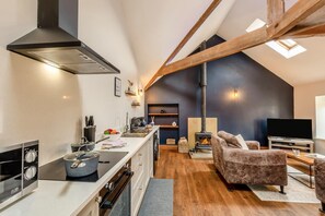 Une cuisine moderne équipée d’une cuisinière, d’un évier et d’une hotte noire. Le plafond est en bois, avec des poutres apparentes. On y trouve également une cheminée et un téléviseur dans un salon confortable, agrémenté d’un canapé et d’une table basse.