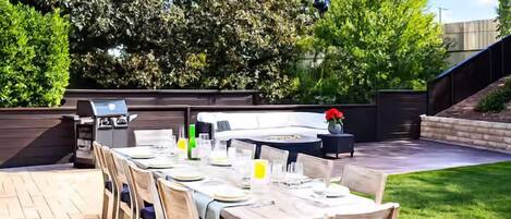 An outdoor dining area with a long table set for four, surrounded by chairs with cushions. There is a grill station to the left and a small table with a vase of flowers to the right.