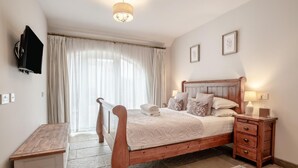 Quarto com cama de madeira, mesas de cabeceira, escrivaninha, TV fixada na parede e uma grande janela com cortinas.