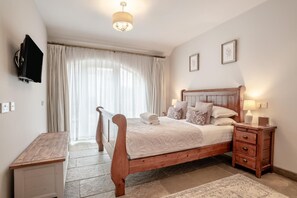 Une chambre à coucher avec un lit en bois, des tables de chevet, un bureau, un téléviseur fixé au mur et une grande fenêtre avec des rideaux.