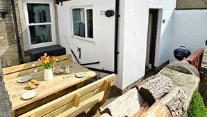 Ein Holztisch mit Tellern und einer Blumenvase, eine Bank sowie ein weißes Gebäude mit Fenstern und einer Tür.