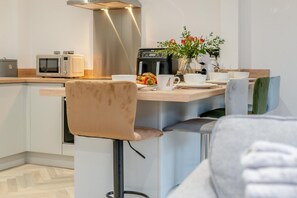 A modern kitchen with a built-in oven, microwave, and coffee machine. There is a bar area with stools and a vase of flowers. - 2 Bed in Padstow (oc-t30497) (Padstow)