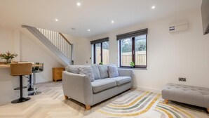 A modern living room with a grey sofa, a dining area with a bar stool, a staircase, and a large window with a view of the outside. - 2 Bed in Padstow (oc-t30497) (Padstow)