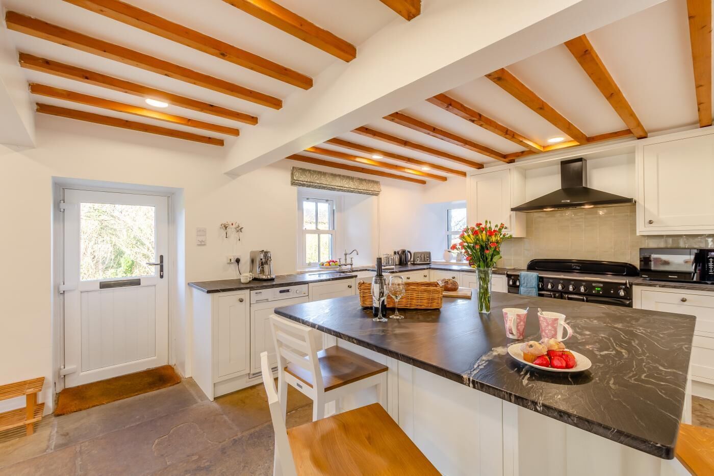 Une cuisine moderne avec des meubles blancs, un îlot en marbre, des poutres en bois au plafond et une grande fenêtre.