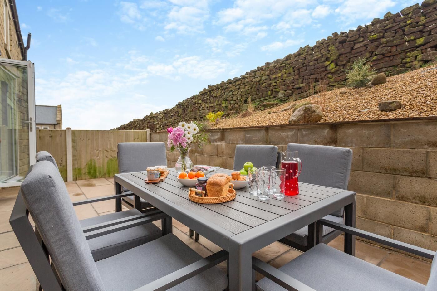 A patio with a table set for breakfast, including bread, fruit, and drinks.