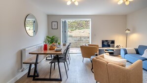 En moderne stue med sofa, spisebord og tv. Et stort vindue giver naturligt lys og udsigt til det fri.
