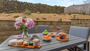 En udendørs terrasse med et bord dækket til et måltid, inklusive forskellige retter og drikkevarer, med en stenvæg og et træhegn i baggrunden.