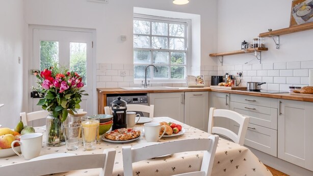A modern kitchen with white cabinetry, a dining table set for breakfast, and a window with a view of trees. - 3 Bed in Brighstone (oc-r30510) (Brighstone)
