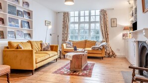 A living room with two sofas, a wooden coffee table, and a fireplace. There are shelves with framed pictures and a large window with curtains. - 3 Bed in Brighstone (oc-r30510) (Brighstone)