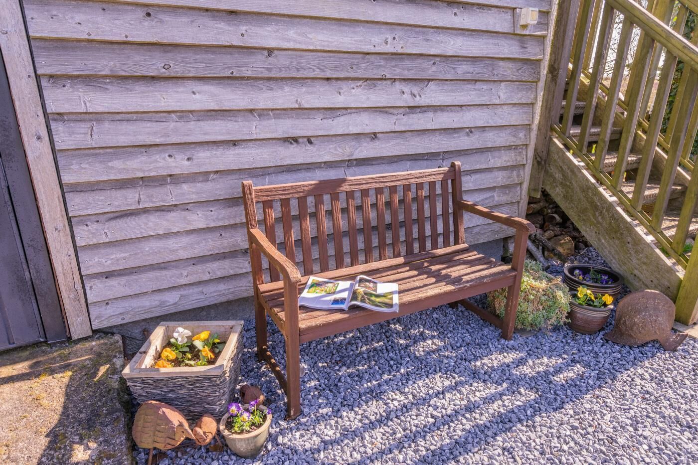 A wooden bench with a magazine, surrounded by gravel, potted plants, and a wooden fence.