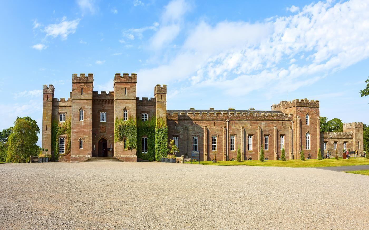 A large, historic stone building with multiple towers and arched windows, surrounded by a gravel courtyard.