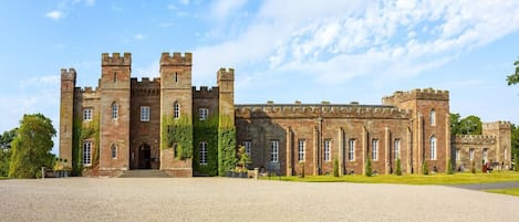 A large, historic stone building with multiple towers and arched windows, surrounded by a gravel courtyard.