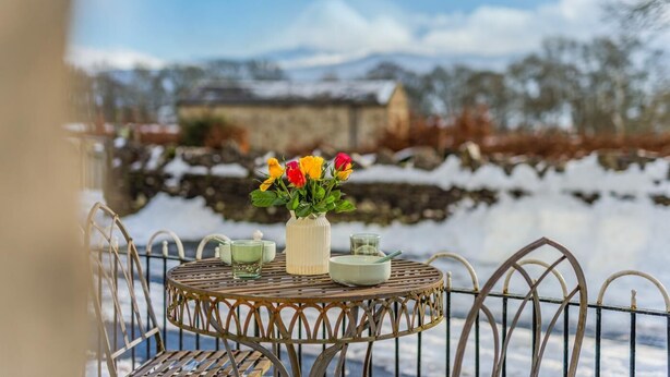 A round metal table with a vase of flowers, a teapot, and two cups on a balcony overlooking a snowy landscape with buildings in the background. - 2 Bed in Hawes (oc-b30521) (High Abbotside)