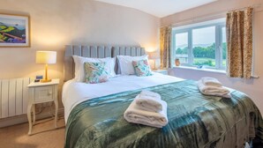 A neatly made bed with a green bedspread, white pillows, and a headboard. There is a bedside table with a lamp and a small vase. A window with curtains is visible, showing a view of greenery outside. - 2 Bed in Hawes (oc-b30521) (High Abbotside)