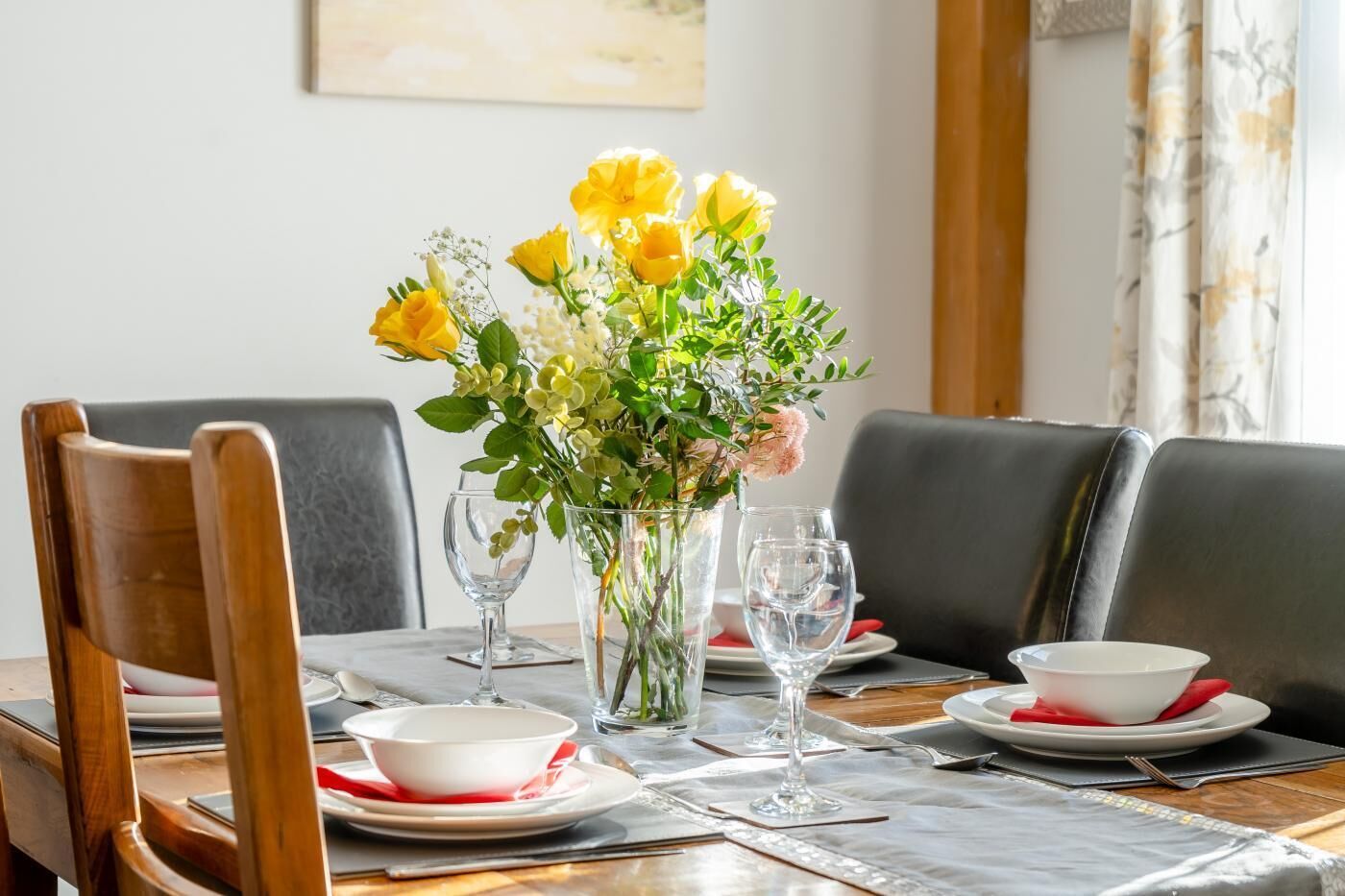 Ein Esstisch ist gedeckt mit weißen Tellern, roten Servietten und klarem Glas. Zwei Vasen mit gelben Blumen und Grünpflanzen stehen darauf.
