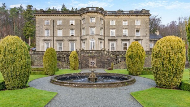 Um edifício grande e histórico com fachada simétrica, uma fonte central e sebes bem cuidadas.
