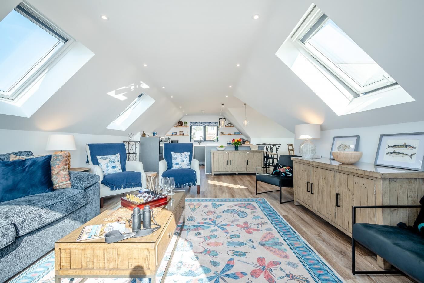 Eine geräumige Loftsage mit hohen Decken, Dachfenstern, einem gemütlichen Wohnbereich mit Sofa und Couchtisch, einer Küche mit Holzausstattung und einem Essbereich mit Stühlen.