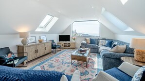 A spacious loft apartment with a high ceiling, skylights, a cozy seating area with a sofa and armchairs, a wooden entertainment unit, and a large window offering a view of the city. - 4 Bed in Porthleven (oc-ch30536) (Porthleven)