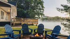 A lakeside deck with Adirondack chairs and a fire pit, overlooking a misty lake and forested hills. - Waterfront Cabin with expansive deck near Saratoga & Lake George (Corinth)