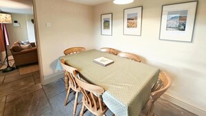 A dining area with a table covered by a green tablecloth, surrounded by wooden chairs. There are framed pictures on the wall, a lamp hanging from the ceiling, and a sofa in the background. - 3 Bed in Garsdale (oc-l30314) (High Abbotside)