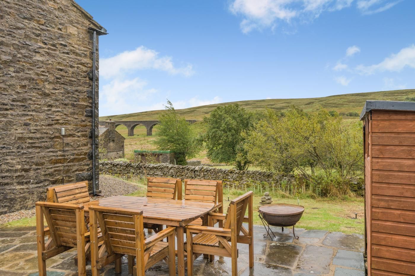 A stone building with a wooden table and chairs, a stone wall, and a view of a bridge in the background.