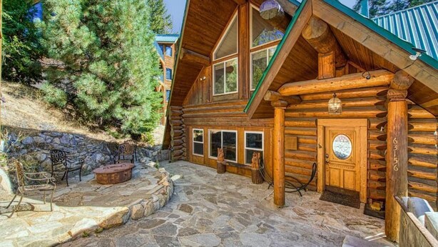 A log cabin with a stone patio, wooden deck, and a large pine tree nearby. - Regatta Chalet – Premium Lakeshore Retreat (Lakeshore)