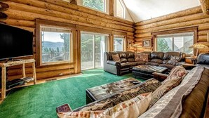 A log cabin interior with a large sectional sofa, a flat-screen TV, and large windows offering a view of the outdoors. - Regatta Chalet – Premium Lakeshore Retreat (Lakeshore)