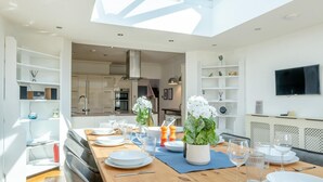 Un comedor moderno con una mesa de madera lista para una comida, una cocina al fondo y un tragaluz arriba.