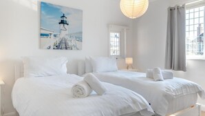Habitación de hotel con dos camas, un cuadro de un faro, una lámpara colgante y una ventana con cortinas.