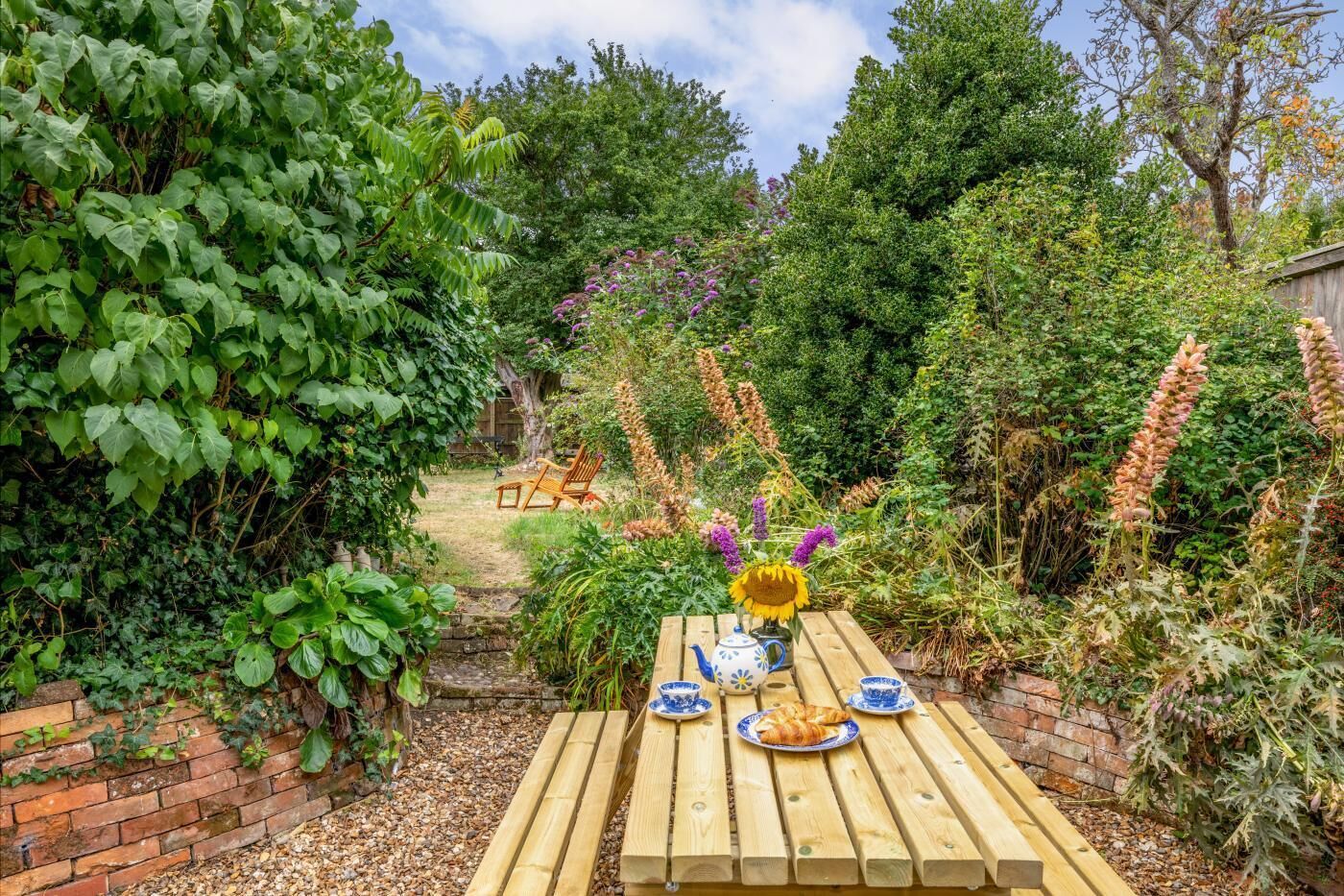 A wooden table set with a teapot, cups, and a plate of food, surrounded by lush greenery and a brick wall.