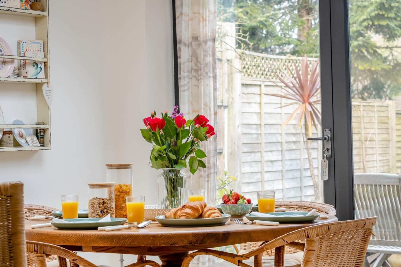 A breakfast table with croissants, fruit, and orange juice, a vase with red flowers, and a wicker chair.