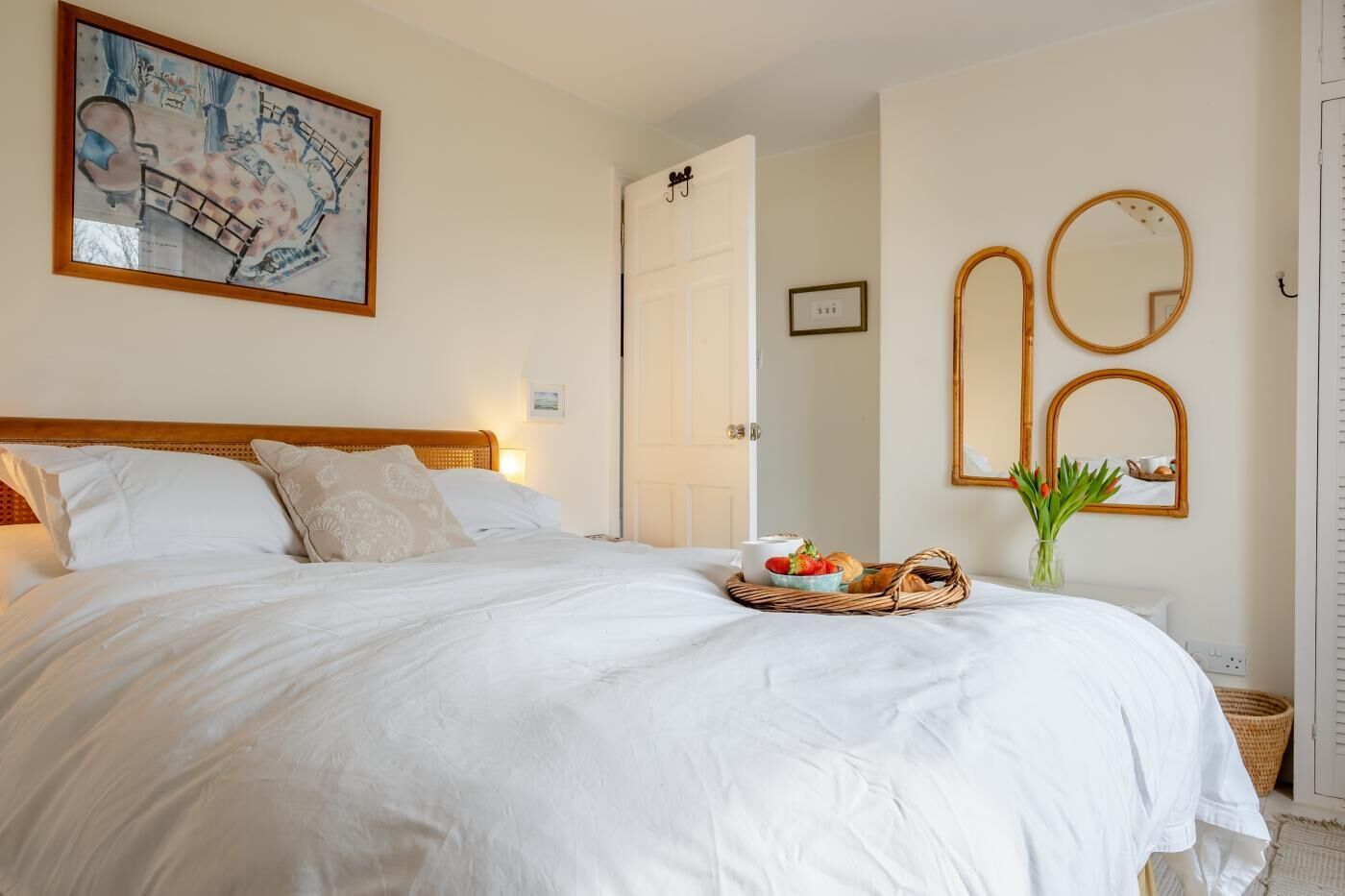 A neatly made bed with white linens, a bedside table with a basket of breakfast items, and a large framed painting above the bed.