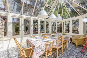 Un hivernacle amb taula de dinar preparada per a sis persones, envoltat de cadires, i una cadira de vimet en un racó.