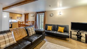 Un salon avec un canapé en cuir noir, un coin repas avec des meubles en bois, un téléviseur posé sur un meuble et un mur en pierre.